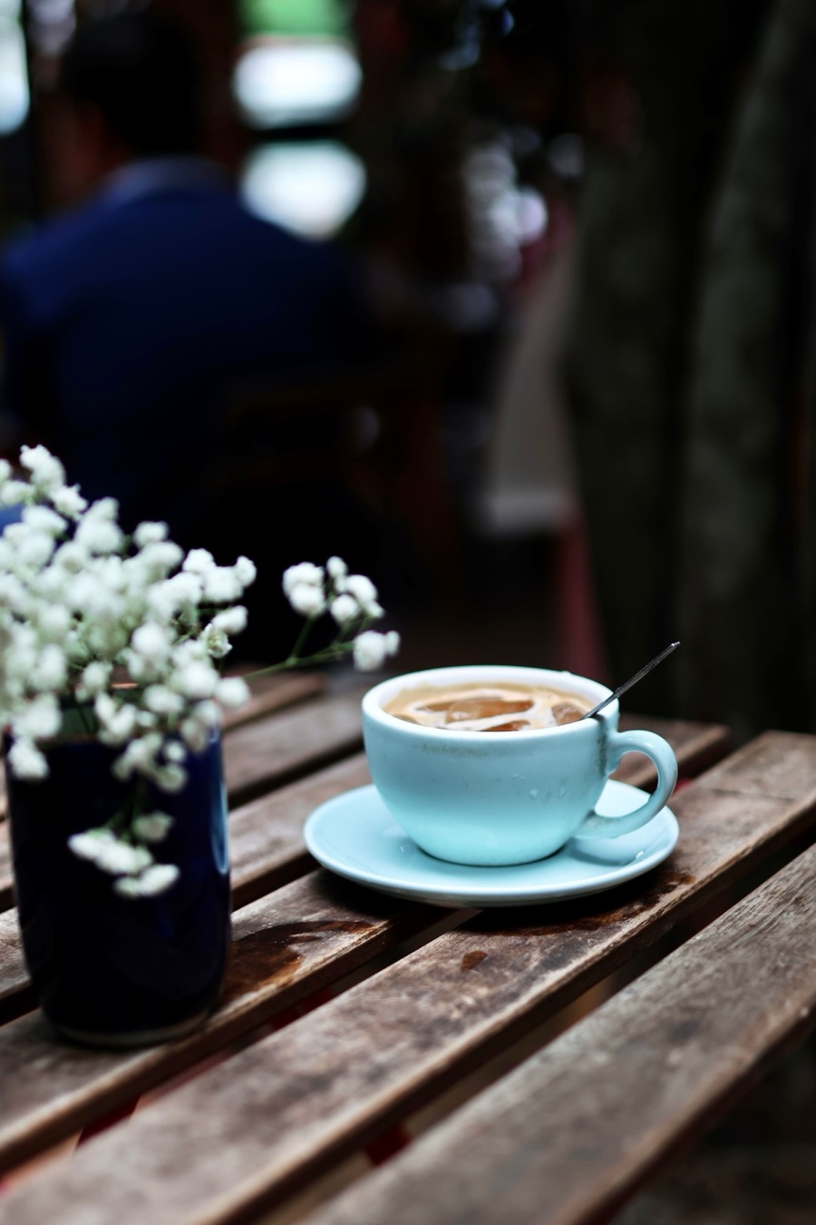 A cup of coffee and a small vase of white flowers on a wooden café table